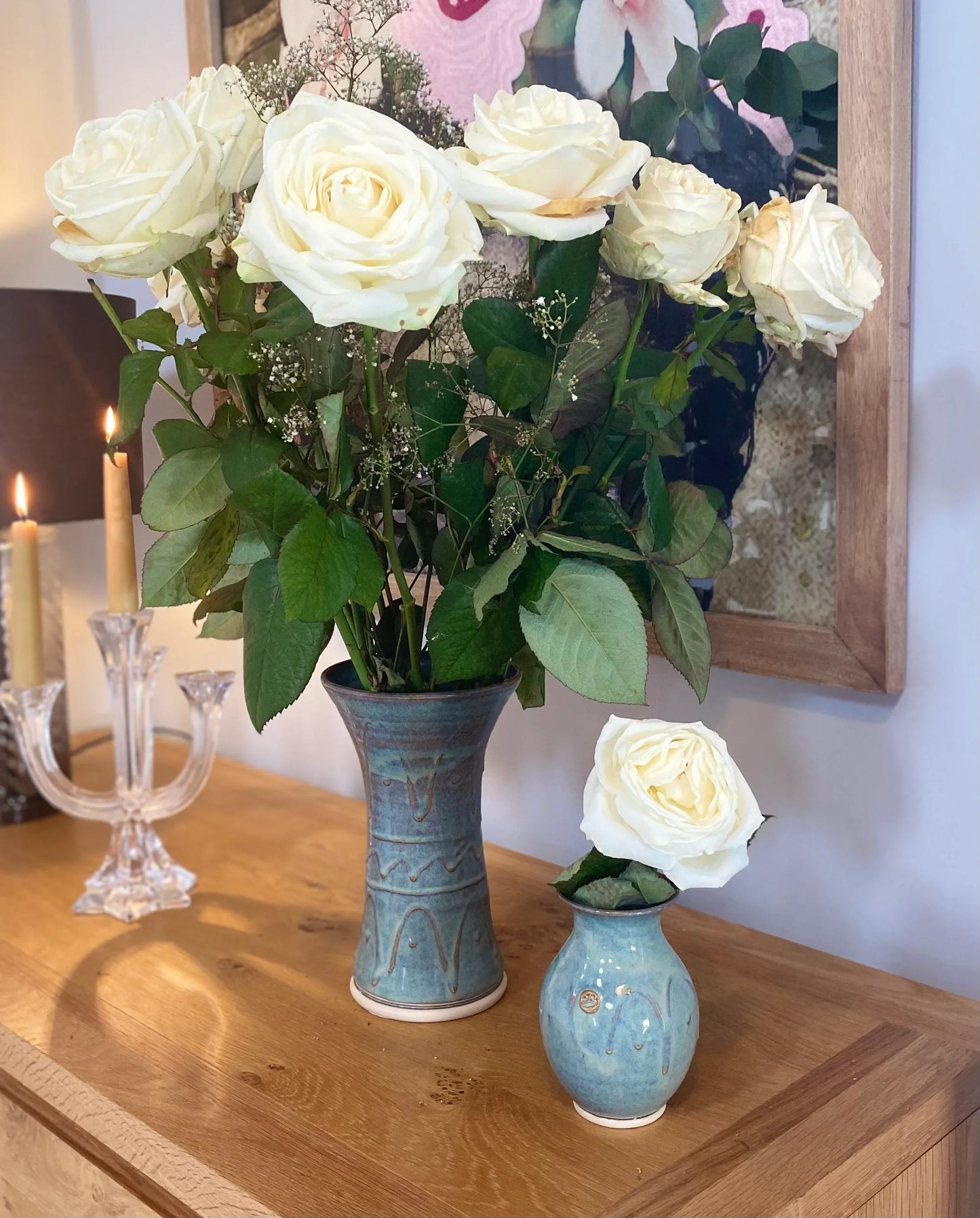 White roses arranged in two handcrafted blue pottery vases on wooden table with lit candles and framed wall art