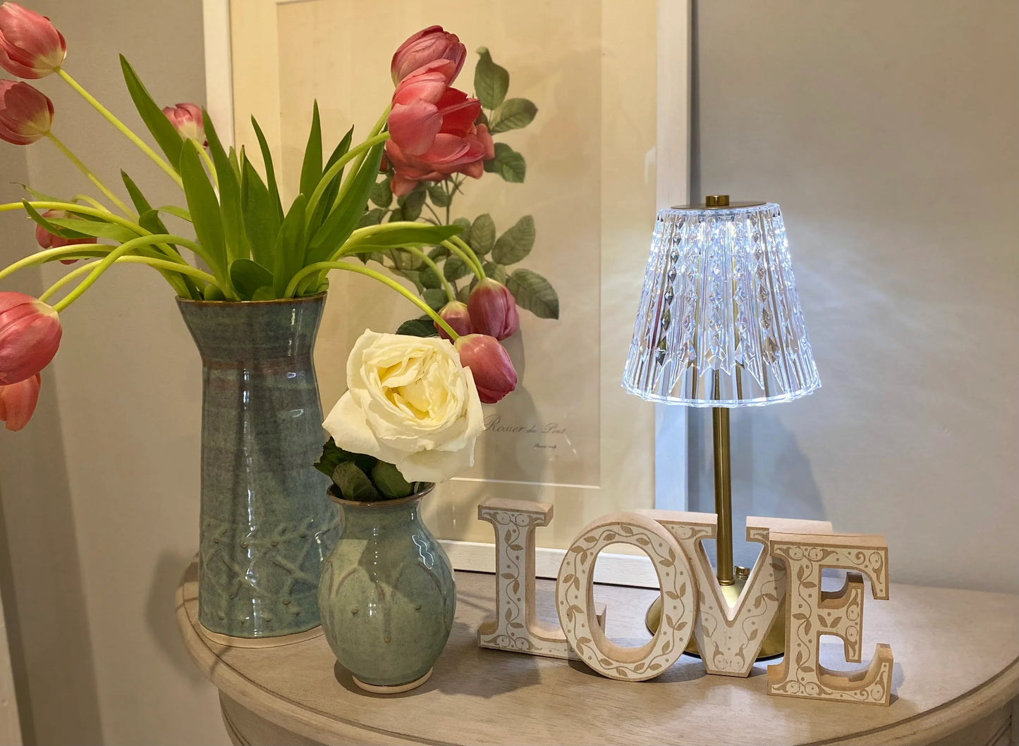 Ceramic vases with pink tulips and a white rose on table beside ornate LOVE sign and modern lamp