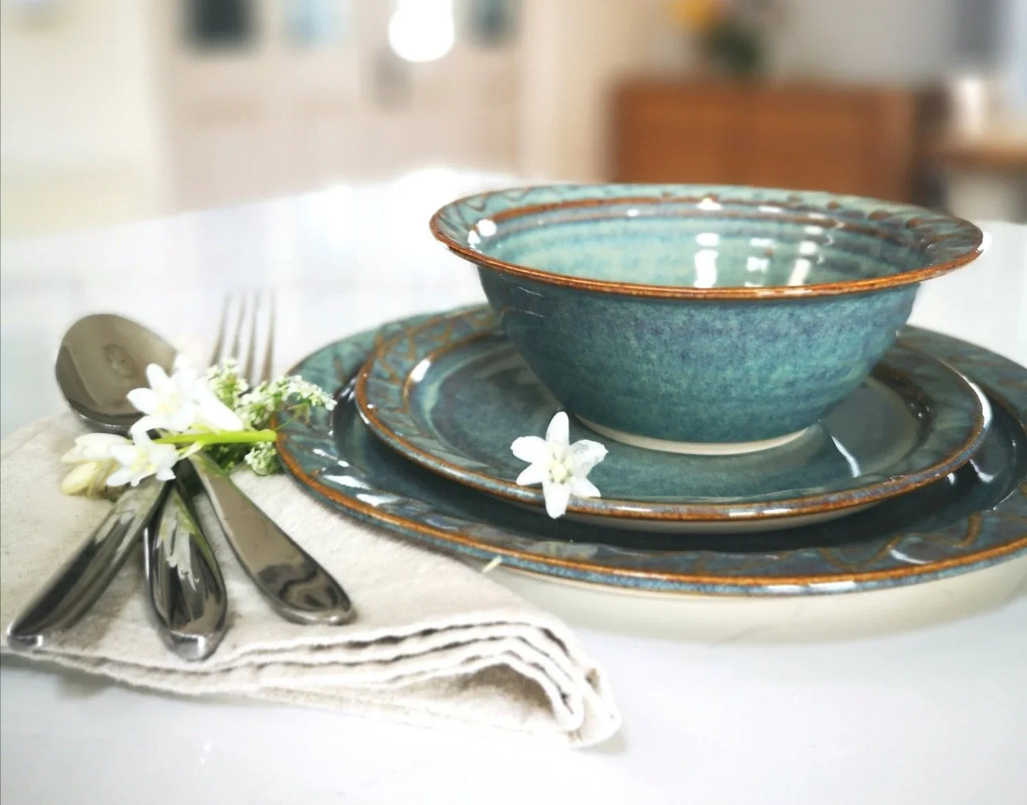 Handcrafted blue ceramic bowl and plates with brown rims on a white table, accompanied by silver cutlery and white flowers
