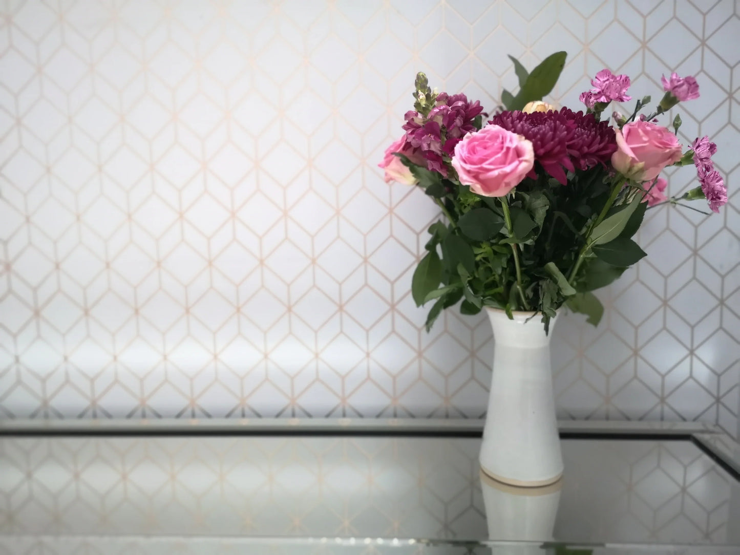 White ceramic vase with pink and purple fresh flowers on reflective glass table against geometric wallpaper