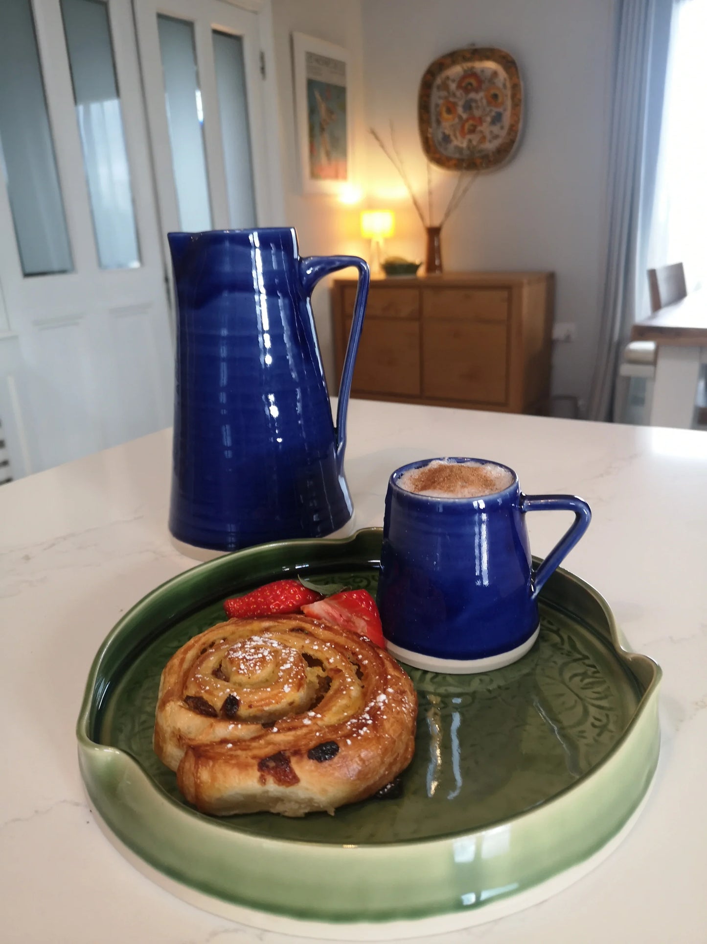 Blue ceramic pitcher and small cup with coffee on green decorative pottery plate with pastry and strawberries on kitchen table