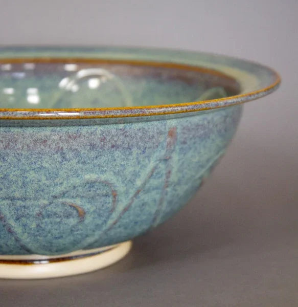 Close-up of blue-green glazed ceramic pottery bowl with subtle embossed floral designs and golden rim