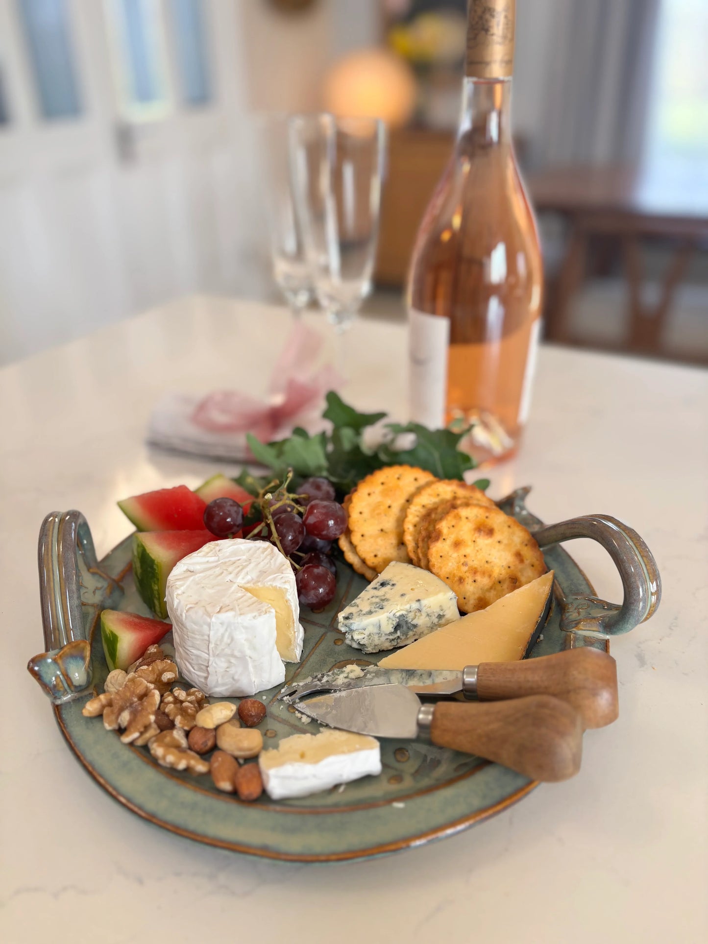 Green ceramic serving plate with assorted cheeses, crackers, nuts, grapes, watermelon, and a bottle of rose wine on a white table