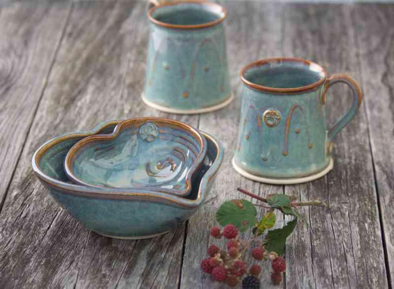 Handcrafted blue-green heart-shaped bowls and two matching mugs on rustic wooden surface, Kilkenny pottery