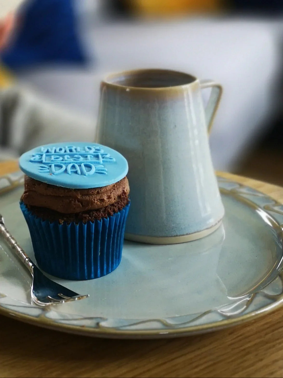 Blue cupcake with chocolate frosting and 'World's Best Dad' topper beside handmade ceramic cup on a glazed ceramic plate