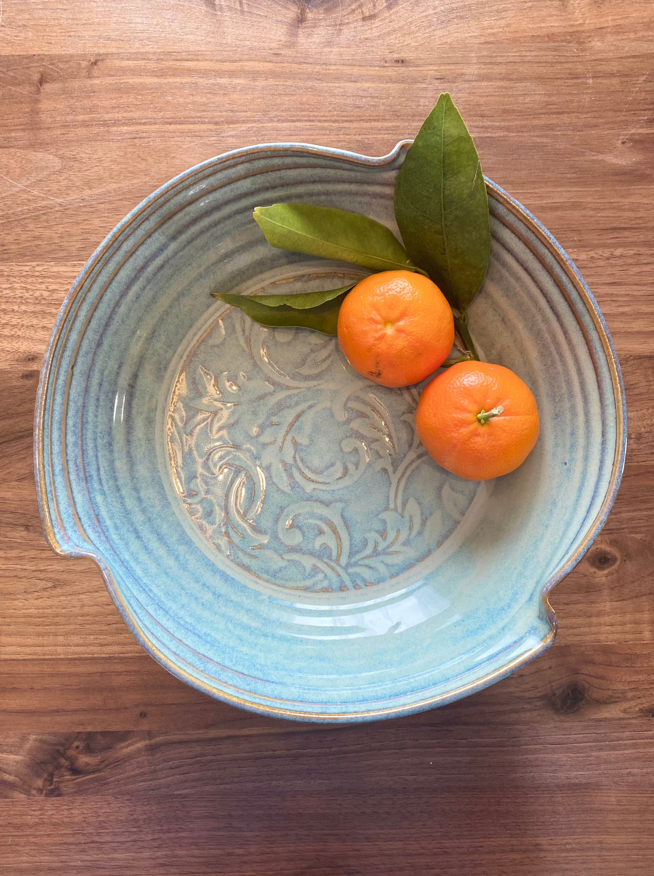Blue ceramic pottery bowl with intricate patterns holding two fresh oranges with green leaves on a wooden table
